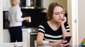 A young woman looks thoughtfully at her phone while sitting at a kitchen table.